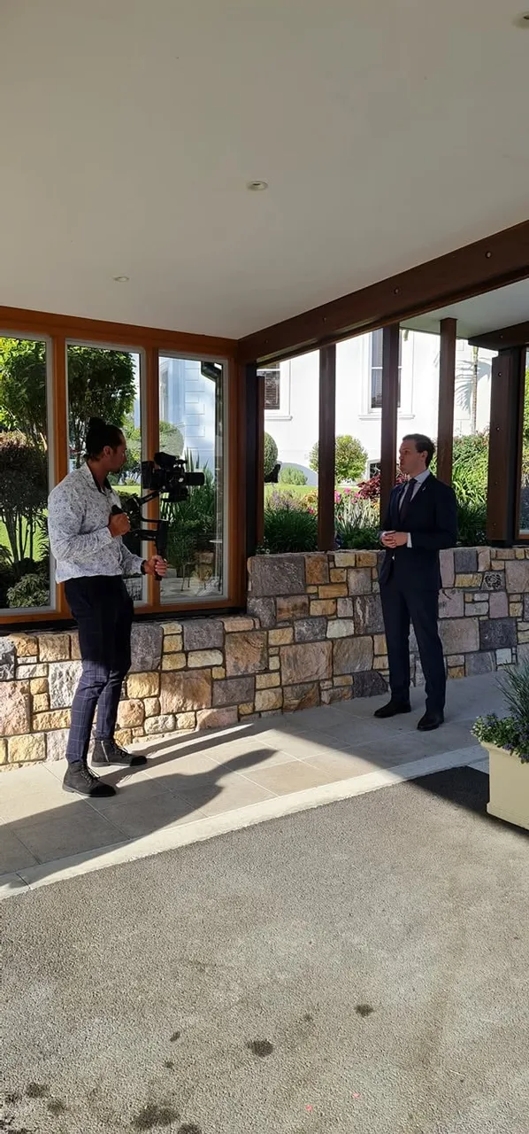 A camera operator filming a presenter beside the windows at Government House